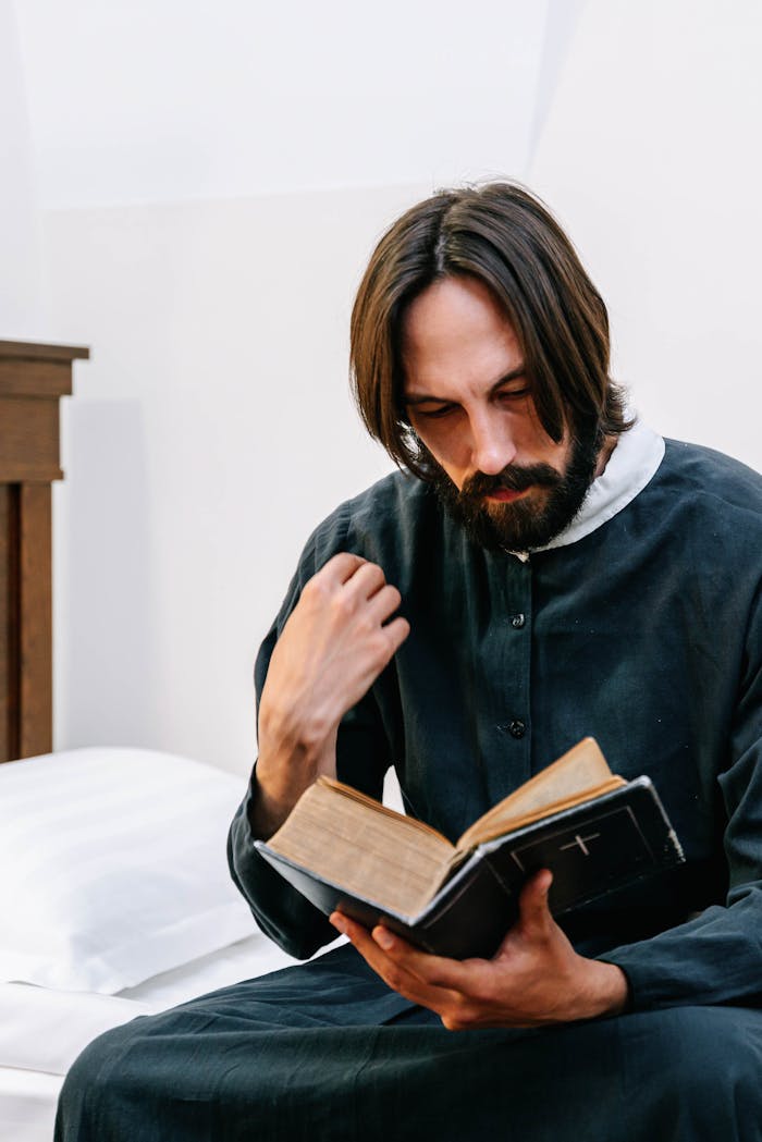 A man wearing a traditional robe deeply engaged in reading a book indoors.